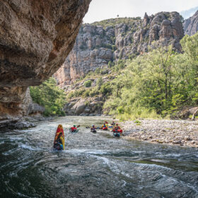 Group of kayakers paddling down a narrow river through a canyon with rocky cliffs and green trees on the banks.