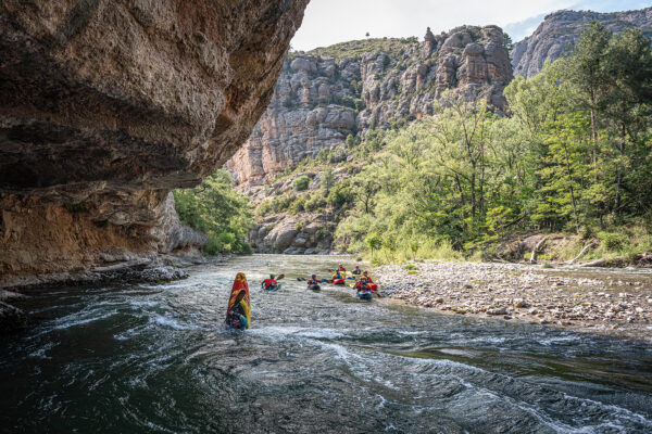 Group of kayakers paddling down a narrow river through a canyon with rocky cliffs and green trees on the banks.